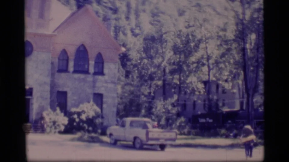 1977 Alaska 8mm frame with the stone building partly framed by trees, a pickup truck, a dark sign, and a person walking at right.