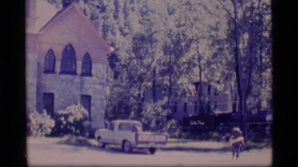1977 Alaska 8mm frame showing the church-like exterior, leafy trees, parked pickup, sign, and a small figure crossing the grass.