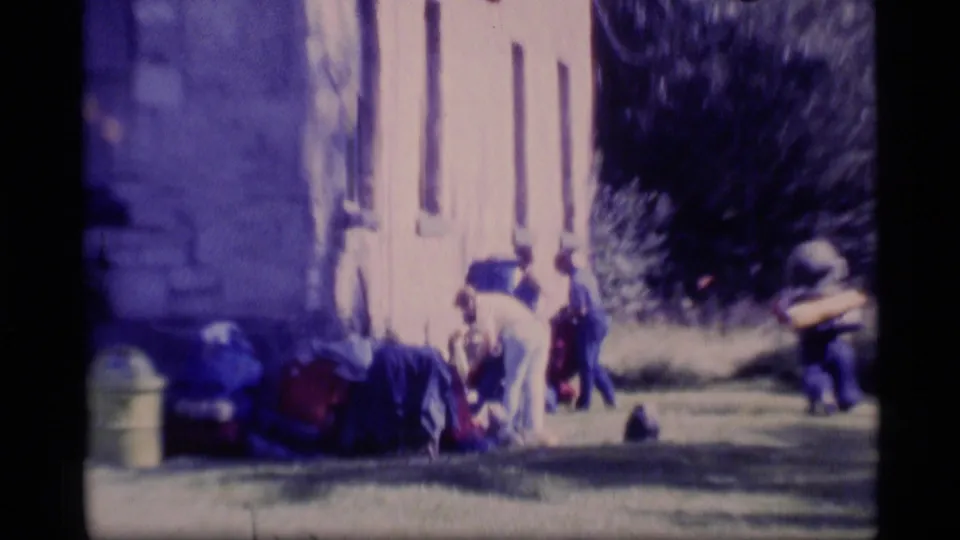 1977 Alaska 8mm frame showing several people gathered beside the church-like building near bags or equipment on the grass.