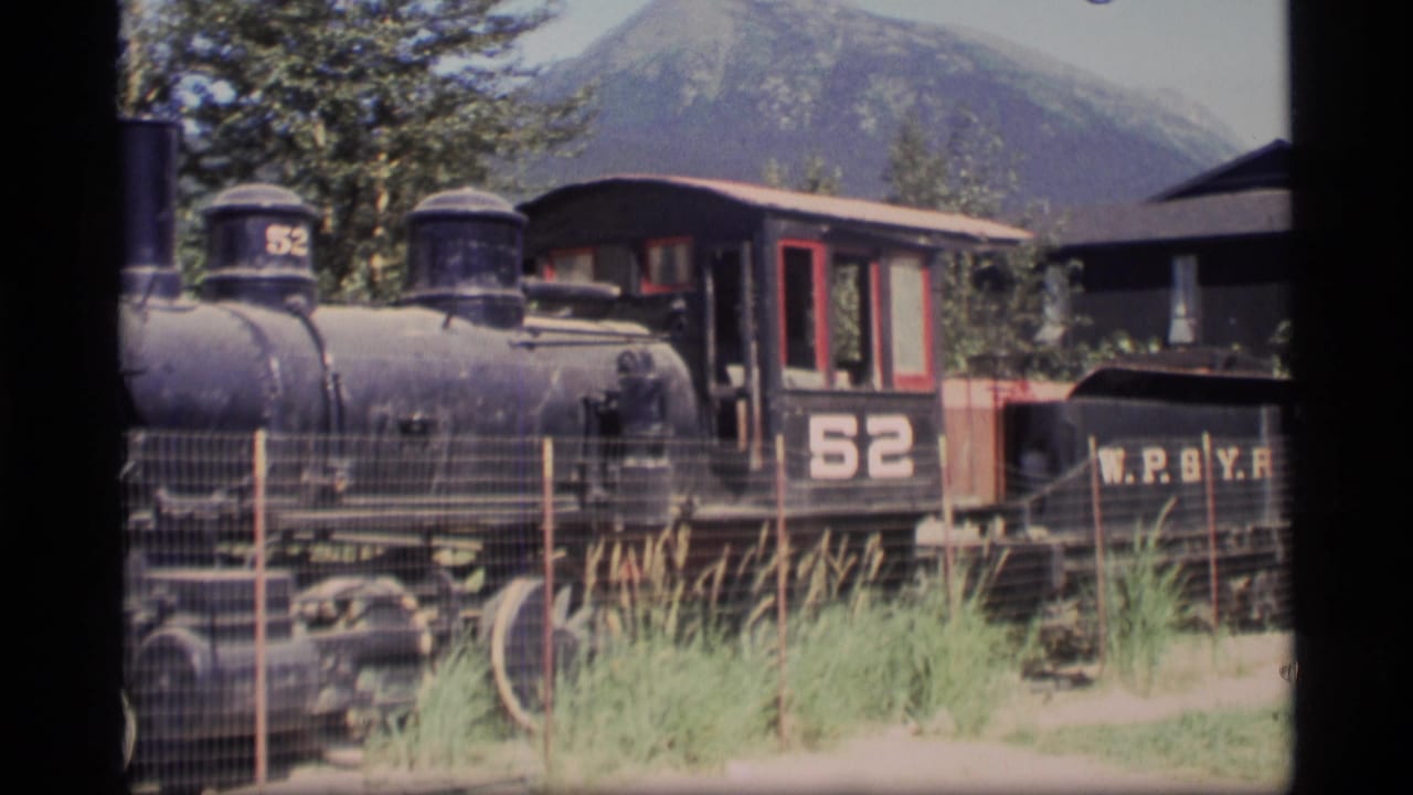 Steam locomotive number 52 behind a wire fence with grass, trees, buildings, and mountains beyond.