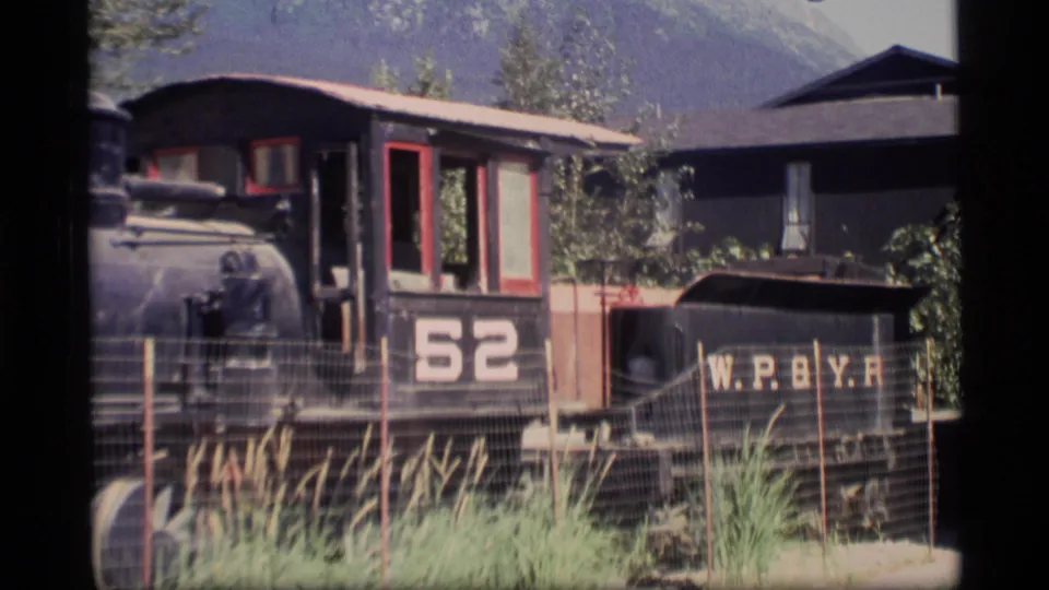 1977 Alaska 8mm frame showing locomotive number 52 and its tender with W.P.B.Y.R. lettering, parked behind a wire fence near buildings and mountain scenery.