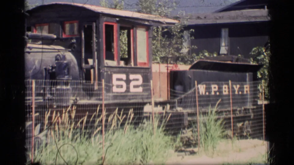 1977 Alaska 8mm frame with a closer look at locomotive number 52, its W.P.B.Y.R.-marked tender, red window trim, wire fencing, and tall foreground grass.