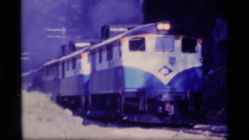 1977 Alaska 8mm frame of a blue-and-white diesel train approaching on the tracks, headlights glowing in a darker, soft-focused rail scene.