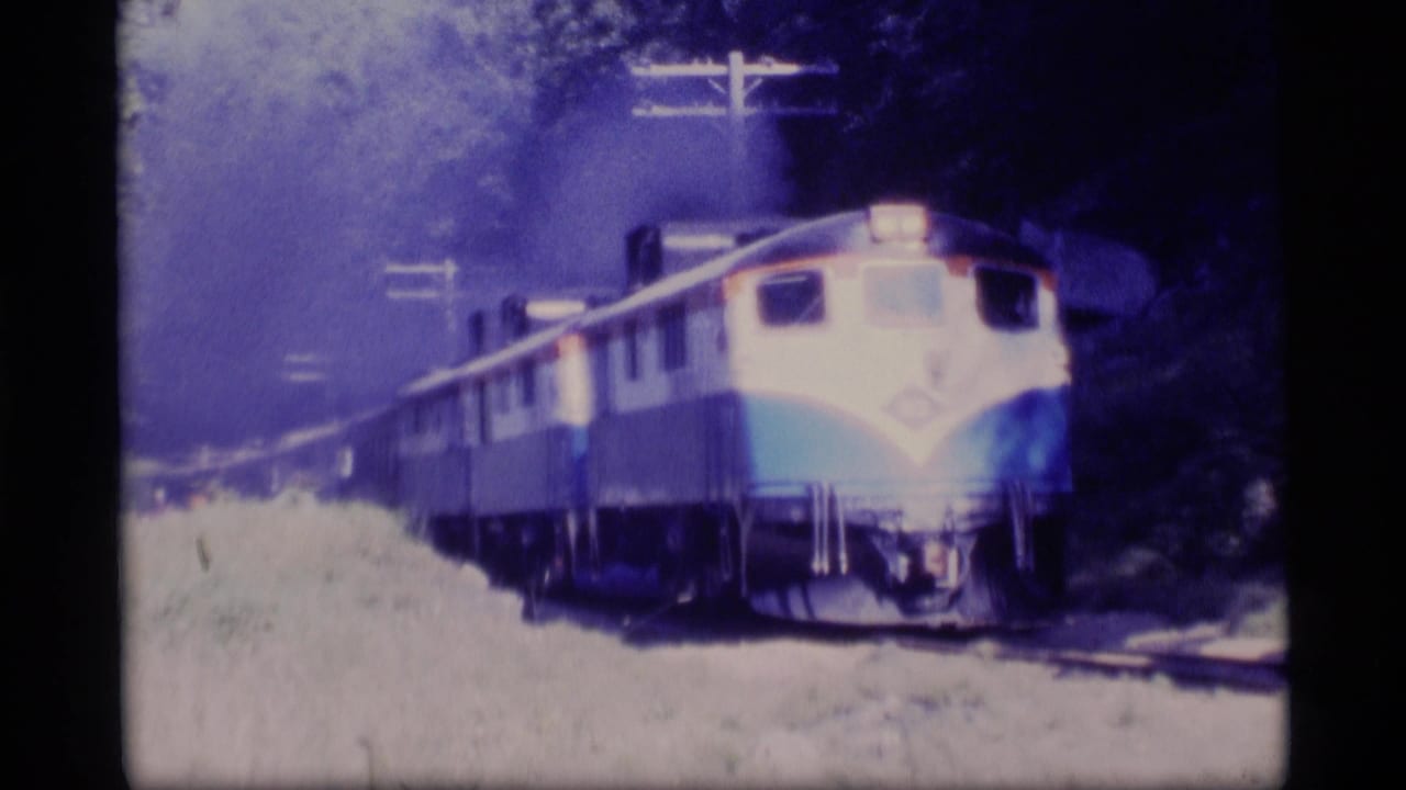 1977 Alaska 8mm frame showing a blue-and-cream passenger train approaching on tracks beside trees and grass.
