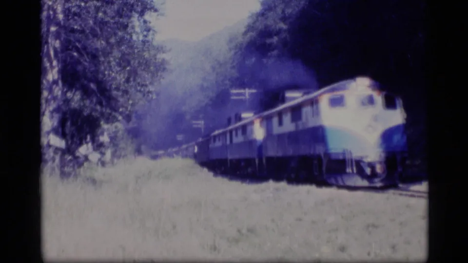 1977 Alaska 8mm frame showing the train farther along the track, framed by trees and a faint hillside.