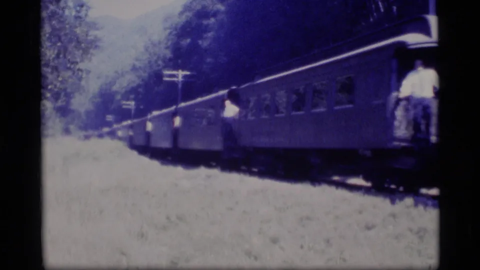 1977 Alaska 8mm frame showing a passenger train beside a wooded landscape, with a rider visible on an open rail-car step.
