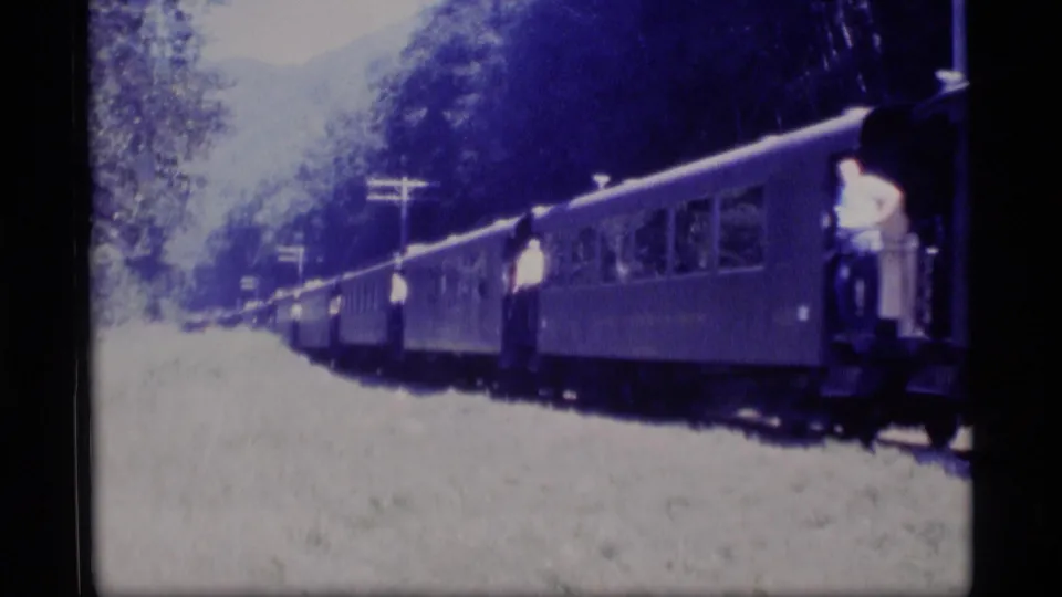 Blue-tinted 8mm view of a passenger train moving through Alaska greenery, with riders visible on open car steps.