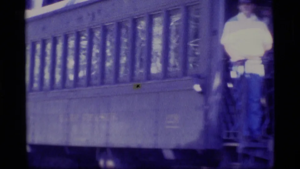 Close 8mm frame of a passenger rail car side with windows and a person standing on the open end step in a white shirt.