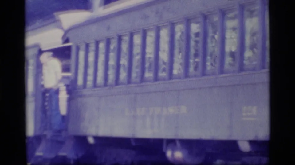 1977 Alaska home-movie frame showing a man in a light shirt standing at the open doorway of a passenger train car.