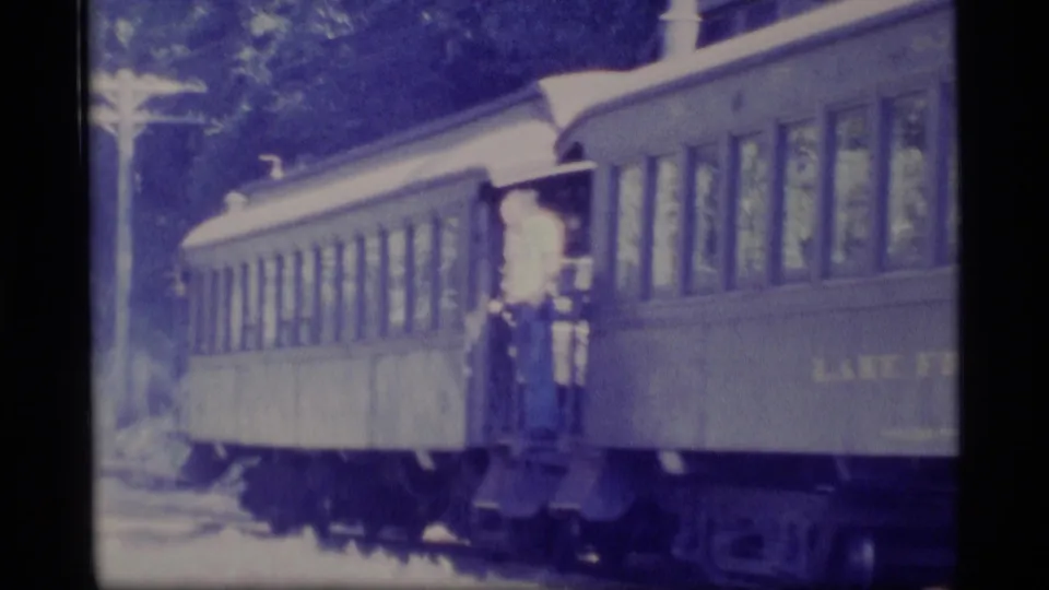 The train car is seen in profile with a man at the vestibule doorway and trees along the trackside background.