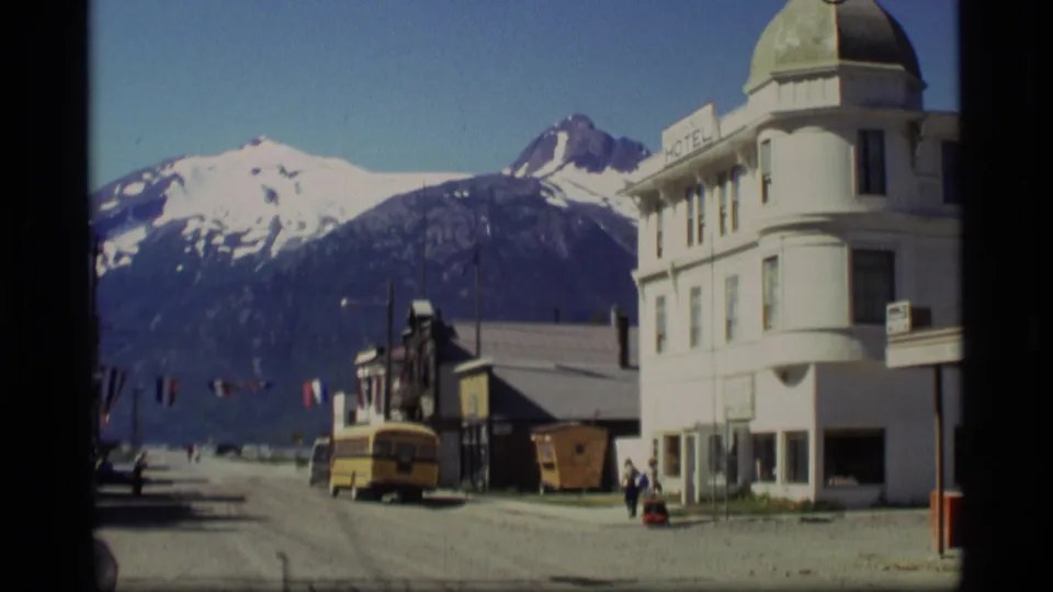 1977 Alaska street view with snow-capped mountains, a white hotel-like building, and a yellow school bus on a gravel road.