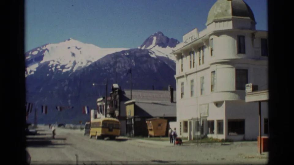 1977 Alaska 8mm frame of a small town street, with pedestrians near a white hotel-like building and mountains behind it.