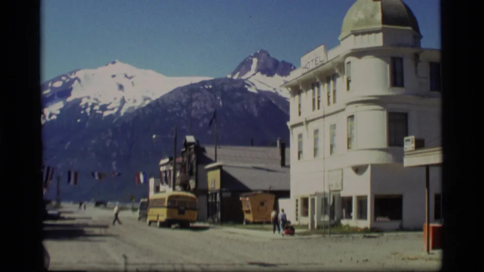 1977 Alaska town street with people walking near a white hotel-like building, a yellow bus, and snow-capped mountains beyond.