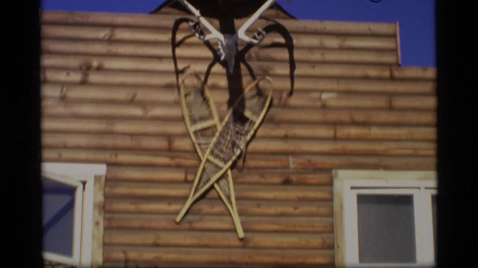 1977 Alaska 8mm frame of a log-sided exterior with a mounted deer skull, antlers, crossed snowshoes, and windows under blue sky.