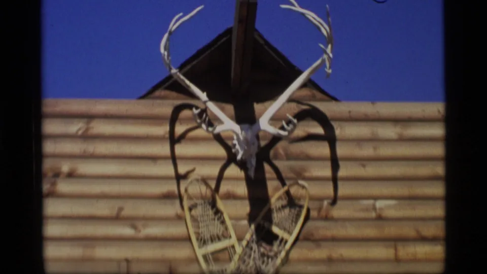Closer 1977 Alaska 8mm view of antlers, skull, and crossed snowshoes mounted on a log wall in bright daylight.