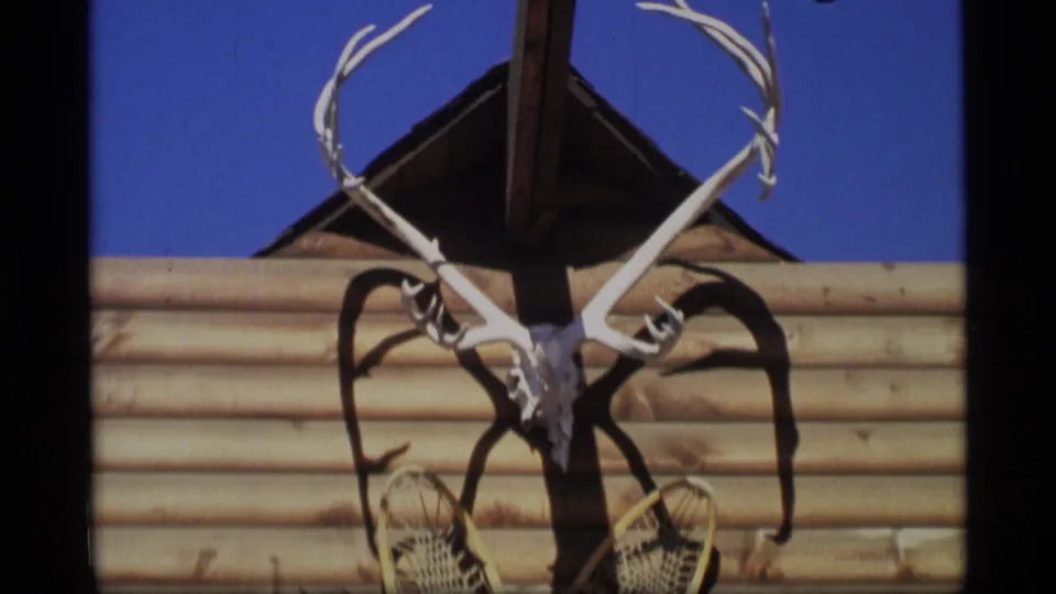 1977 Alaska 8mm frame centering a mounted skull, branching antlers, and crossed snowshoes on a log-sided building.