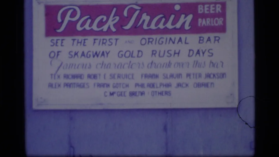 Close 1977 Alaska frame of a Pack Train Beer Parlor sign referencing Skagway Gold Rush days on a blue-painted wall.