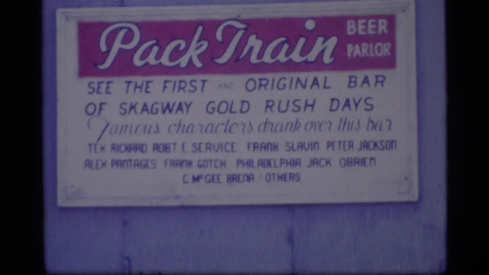 1977 Alaska 8mm close-up of the Pack Train Beer Parlor sign, with painted text about Skagway Gold Rush days and listed names.