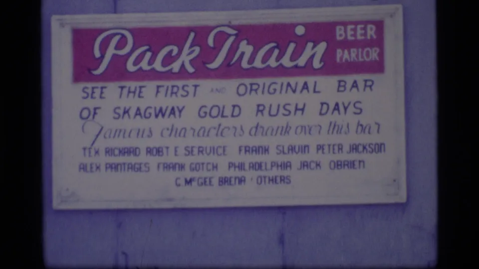 Tight 1977 Alaska home-movie frame of the Pack Train Beer Parlor sign with red header and Gold Rush reference text.