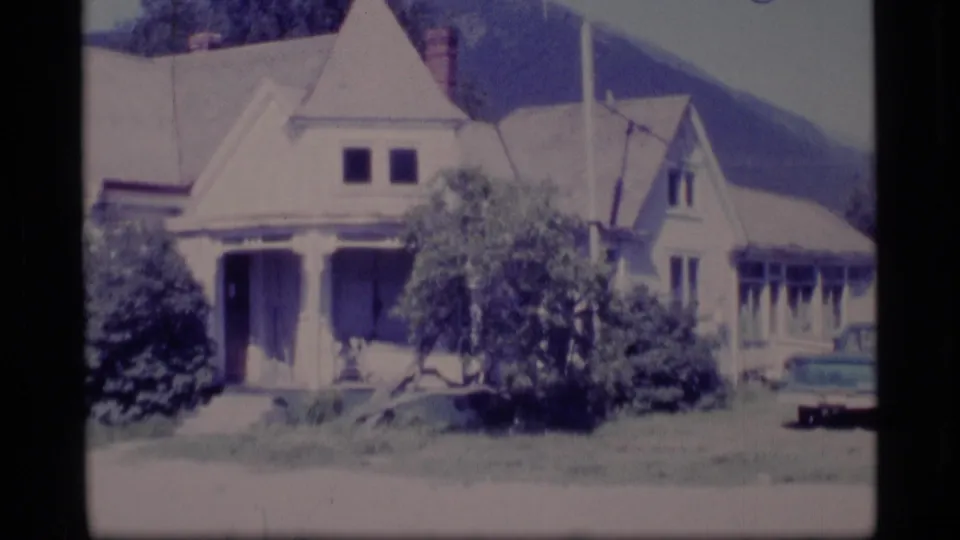 1977 Alaska 8mm frame showing a gabled suburban house exterior with porch, shrubs, lawn, and a car partly visible at the edge.