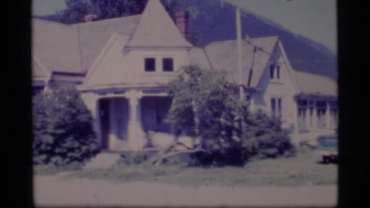 Older light-colored house with porch, shrubs, lawn, and wooded hill behind it.
