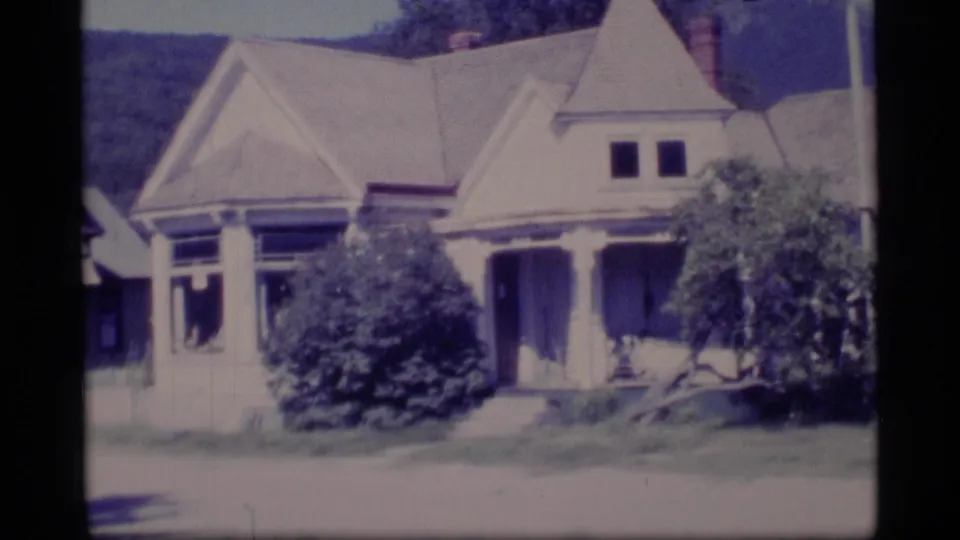 1977 Alaska residential view with a porch-front gabled house, dense shrubs, lawn area, and wooded hill beyond the roofline.