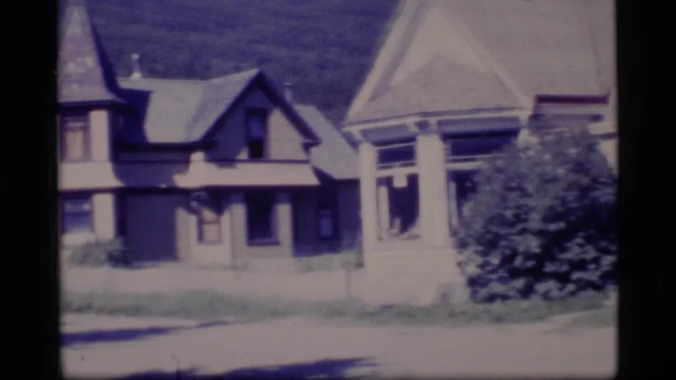 1977 Alaska 8mm frame showing neighboring older houses, steep rooflines, porch columns, shrubs, and a pale road or driveway edge.