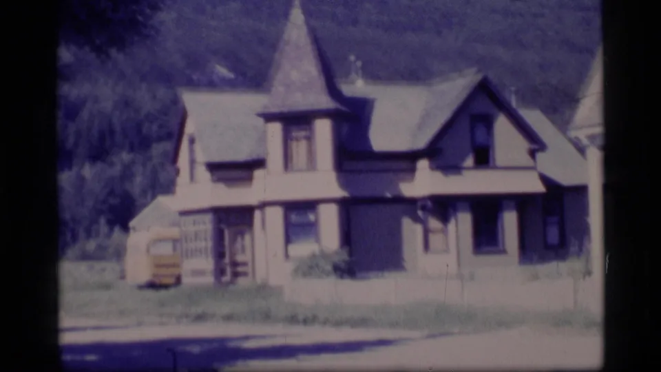 1977 Alaska frame showing a house with a turret-like roof form, a small yellow vehicle nearby, grass, road edge, and wooded hillside.