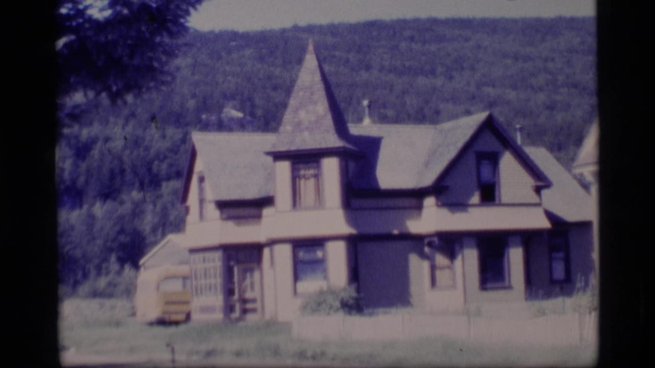 1977 Alaska 8mm frame showing a light-colored old-style house with a pointed turret, trees, and wooded hillside.