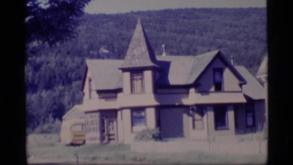 1977 Alaska 8mm frame showing a light-colored old-style house with a pointed turret, trees, and wooded hillside.