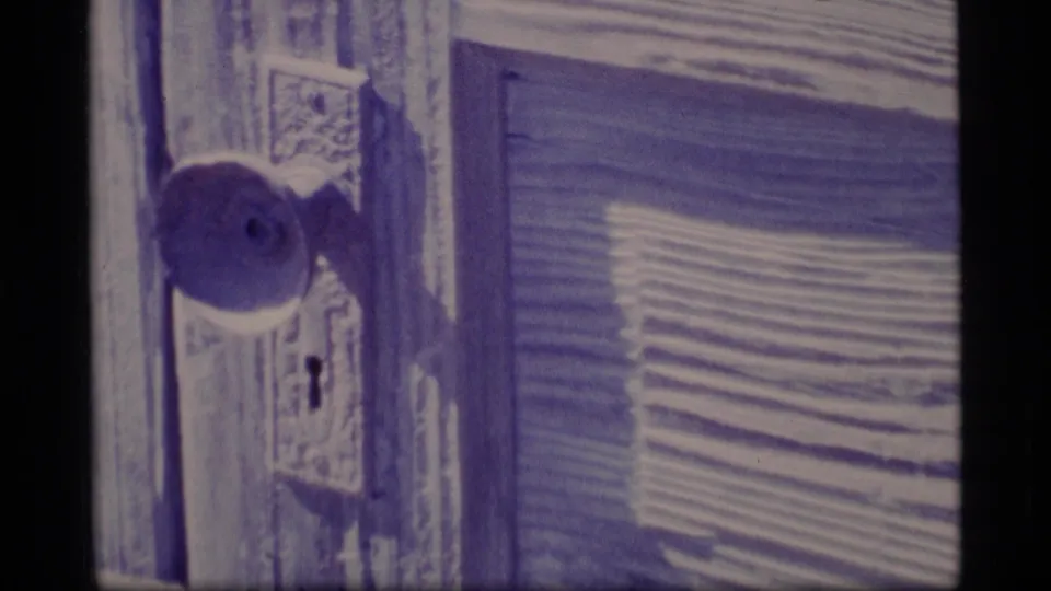 Close 8mm view of an older wooden door, round doorknob, keyhole, and adjacent shuttered siding in Alaska footage from 1977.