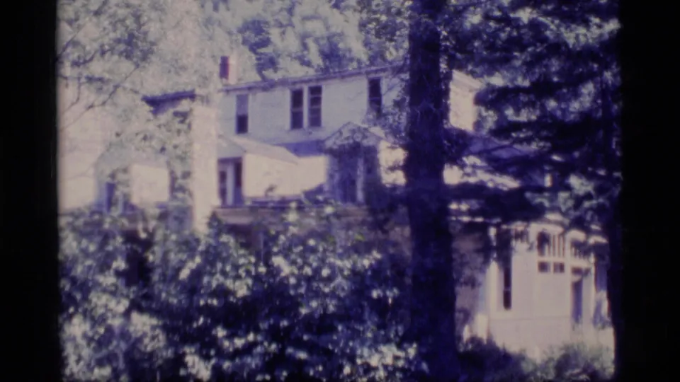 1977 Alaska 8mm frame showing a large pale house partly obscured by leafy shrubs, trees, and a porch or railing.