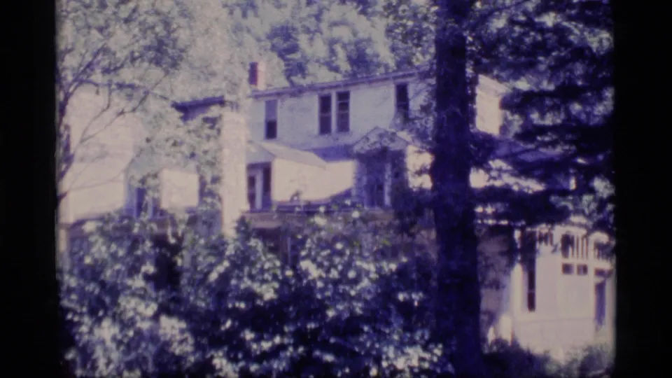 1977 Alaska 8mm frame showing a white house behind dense trees and shrubs, with soft focus and film grain.