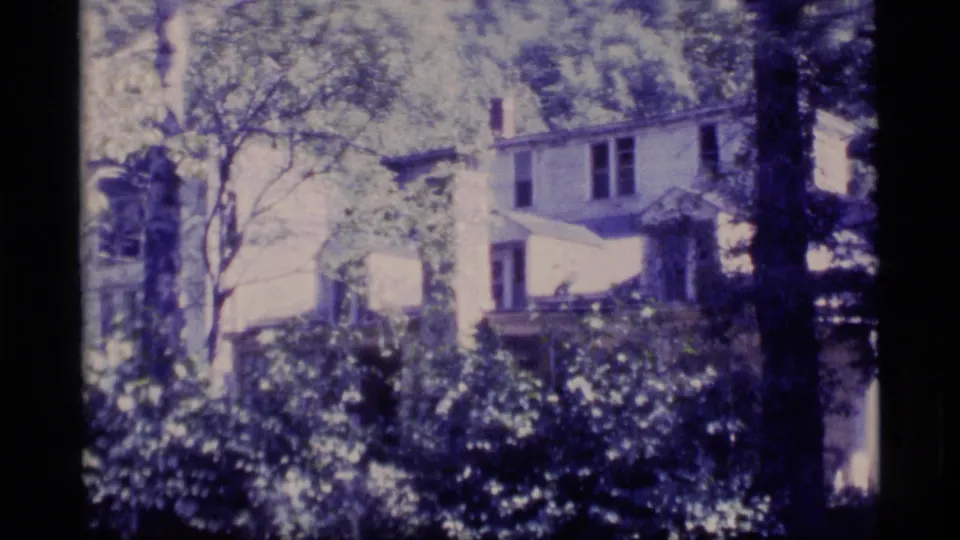 A white residential exterior appears beyond trees and shrubs in grainy 1977 Alaska home-movie footage.