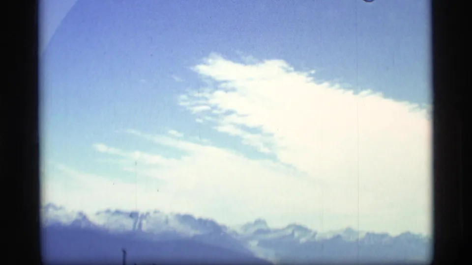 1977 Alaska 8mm view of pale blue sky, bright clouds, and distant snow-capped mountains.