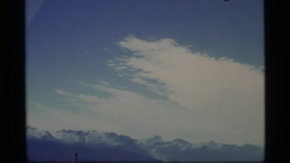 1977 Alaska home-movie frame showing distant snow-capped mountains under a blue sky with wispy clouds.