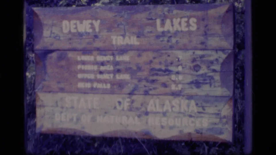 1977 Alaska frame showing a close view of a weathered Dewey Lakes Trail sign with State of Alaska natural resources text.