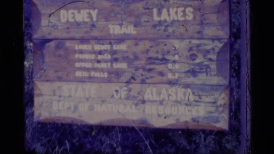 1977 Alaska home-movie frame with the Dewey Lakes Trail sign and partially legible State of Alaska department text.