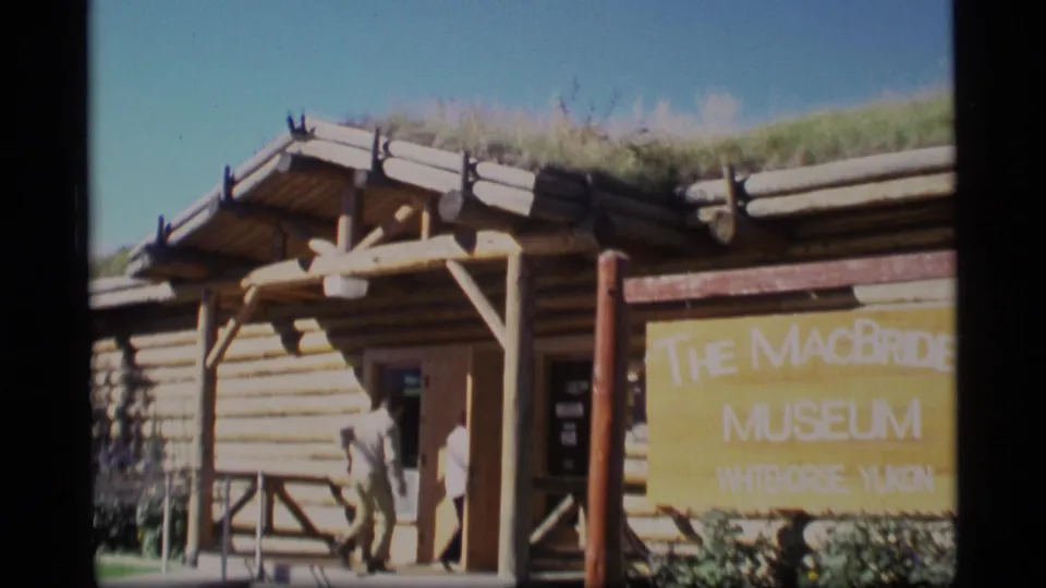 The MacBride Museum exterior in Whitehorse, Yukon, with log walls, a grass roof, yellow signage, and visitors entering or standing near the doorway.