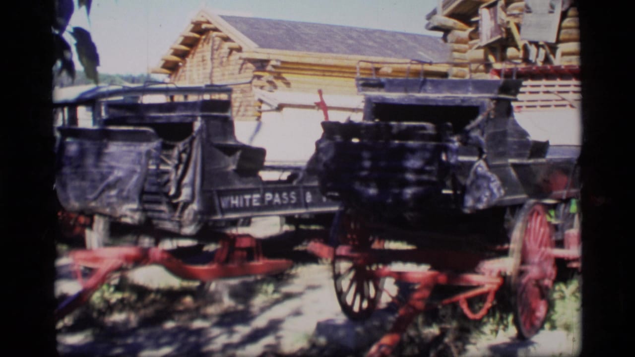 1977 Alaska 8mm frame showing two historic black carriages with red wheels near a log-style building, one partly marked White Pass.