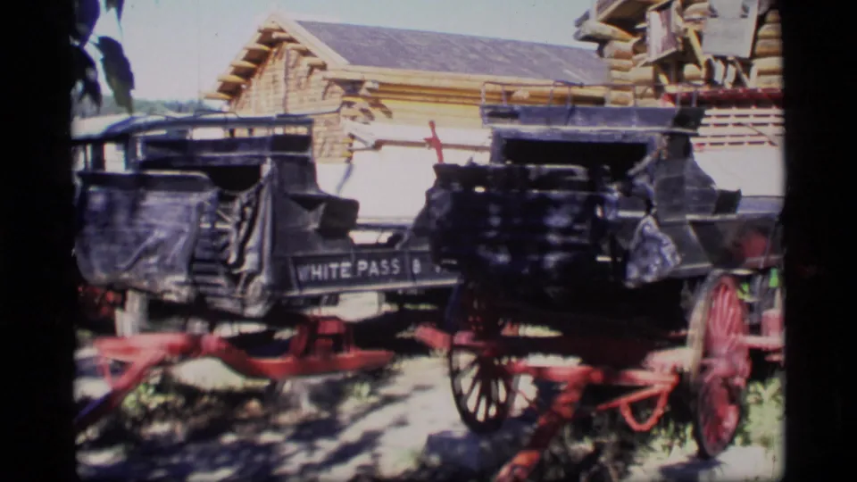 1977 Alaska 8mm frame showing two historic black carriages with red wheels near a log-style building, one partly marked White Pass.