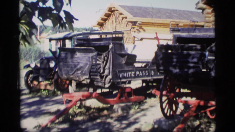 Visitor-shot 1977 Alaska frame of a White Pass carriage display with black bodies, red wheels, leafy shadows, and a rustic building behind.