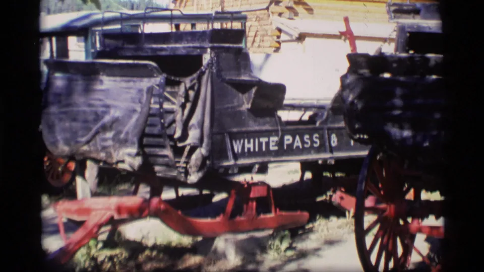 Closer 1977 Alaska 8mm view of historic carriage bodies, red wheel hardware, and visible White Pass lettering outside a rustic structure.