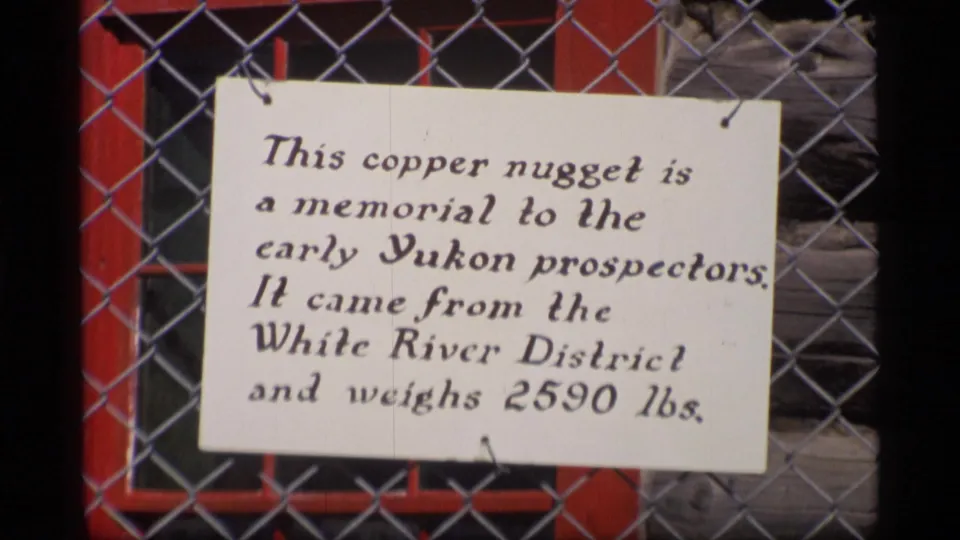 1977 Alaska frame showing a chain-link fenced sign stating that a 2,590 lb copper nugget from the White River District memorializes early Yukon prospectors.