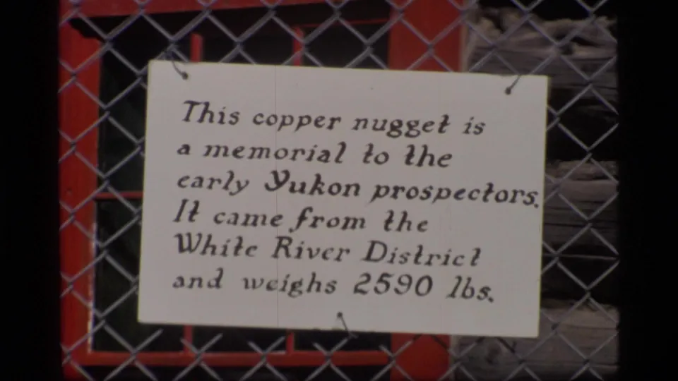 Close view of a handwritten Alaska memorial sign for a copper nugget, filmed through a chain-link fence in 1977.