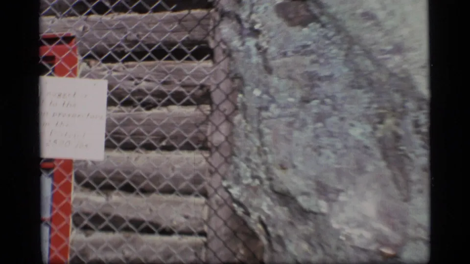 Wider 1977 Alaska view showing the memorial sign at left and a large weathered rock behind chain-link fencing.