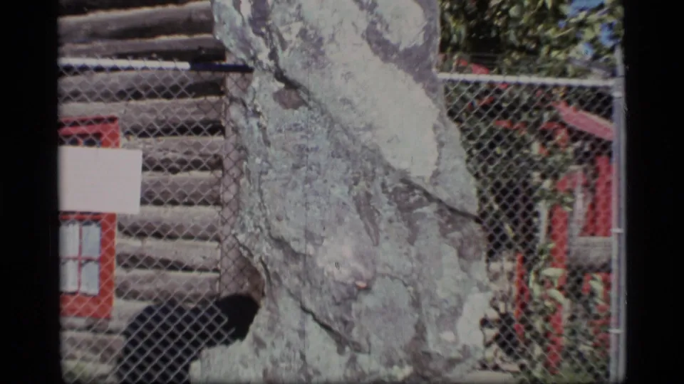 Wider view of the fenced copper nugget display, with red framing, stacked logs, and greenery visible in 1977 Alaska footage.