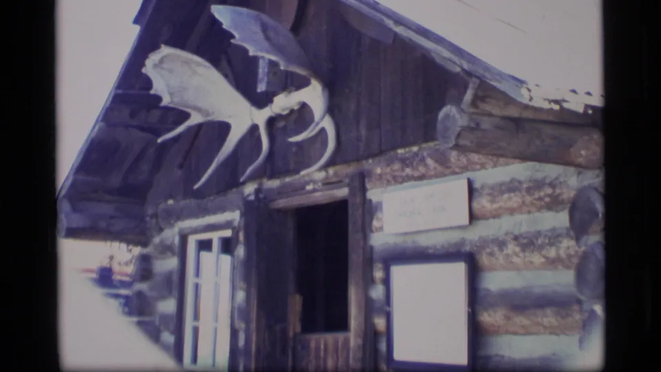 1977 Alaska 8mm frame showing a log cabin entrance, mounted antler display, roof overhang, open doorway, and posted signs.