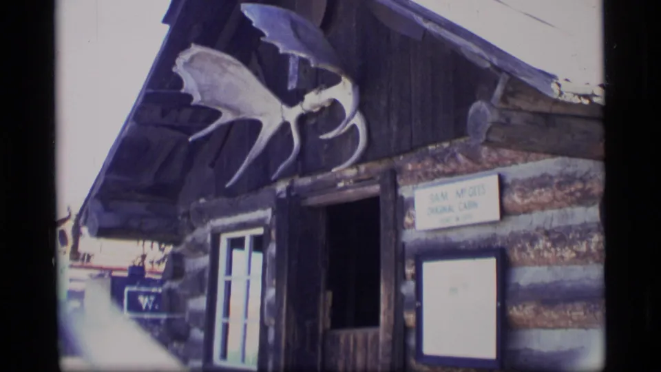 1977 Alaska 8mm frame with the Sam McGee's Original Cabin sign, log facade, entrance area, and mounted antler display.