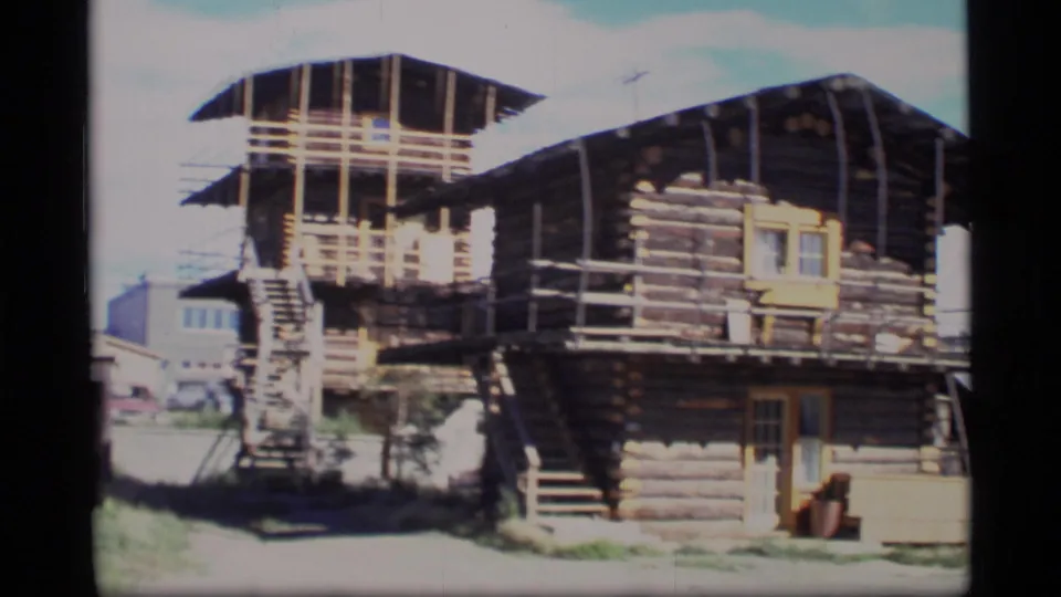 1977 Alaska 8mm frame showing a multi-level log cabin structure with exterior stairs, railings, roofed upper level, and nearby buildings.